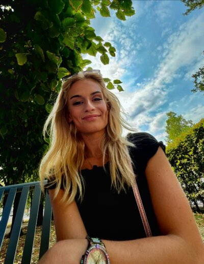 A Czech girl with long blonde hair, wearing a black top and bracelets, is sitting on a bench under a tree