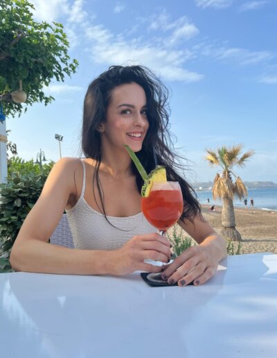 A Czech wife with long dark hair sits at an outdoor table by the beach, holding a red cocktail garnished with a pineapple slice and a green straw